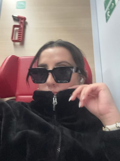 The image displays a female individual inside a public transport vehicle, likely a train or bus, potentially operating in or around Brunn am Gebirge, Austria. The subject is centrally framed from the mid-torso upwards, wearing dark rectangular sunglasses and a black zippered jacket of a fluffy or fleece texture. She has dark hair, a white wireless earbud in her right ear, and a small gold hoop earring in her left. Her left hand, with manicured nails featuring white tips, is raised towards her neck, revealing a white bracelet with a gold-colored clasp and a four-petal flower design on her wrist. Behind her to the left, a red upholstered seat is partially visible. A red emergency hammer is mounted on a light-colored wall panel above the seat, with a light green square sign situated above the hammer. To the right of the subject, a white wall section features a partially obscured green sign.