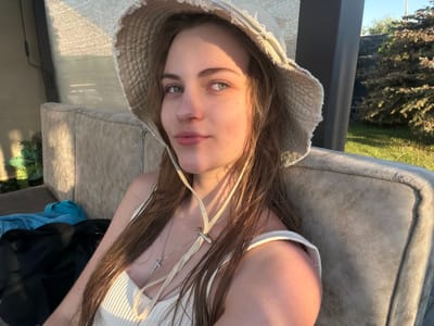 The media file features a young person lying down, wearing a white tank top and a bucket hat. They have long, brown hair and green eyes, and are wearing a silver necklace with a cross pendant. The person is lying on a textured, light-colored surface, possibly an outdoor couch or lounge chair.

The setting appears to be outdoors, with greenery and trees visible in the background, suggesting a daytime scene. The lighting indicates it might be late afternoon or early evening, with sunlight casting shadows and highlighting textures. The overall mood seems relaxed and peaceful, with the subject looking towards the camera with a neutral expression.

There is no visible text in the media file. The location context provided is Slobozhanske, Ukraine.