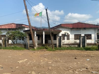 The image displays a daytime scene featuring a single-story white building with a red corrugated metal roof, partially obscured by two prominent wooden utility poles and overhead wires. The building appears weathered, with peeling paint and dirt accumulation on its walls and the surrounding concrete fence. A metal gate is visible in the center of the fence.
On the building's facade, to the right, a rectangular sign with black text on a white background is mounted. The visible text reads, in French: "MINISTERE DE L'ELEVAGE, DE LA PECHE ET DES INDUSTRIES ANIMALES", which translates to "Ministry of Livestock, Fisheries, and Animal Industries". Below this, another line of text is less clear, likely indicating a local delegation. An outdoor air conditioning unit is installed below the sign. The windows of the building are fitted with security shutters or bars.
A flag, displaying horizontal stripes of green, yellow, and red, with a star or emblem (characteristic of the Cameroonian flag), is flying from a pole situated behind the building on the left side. Green foliage, possibly shrubs or small trees, is visible behind the fence on the left.
The foreground consists of a rough, unpaved dirt surface, heavily littered with debris including plastic bags, paper, and other waste. The sky above is bright and clear, with scattered white clouds, suggesting good weather conditions. In the distant background, faint outlines of hills or mountains can be seen. The scene depicts a functional government building in what appears to be a somewhat neglected or rural environment in Victoria, Cameroon.