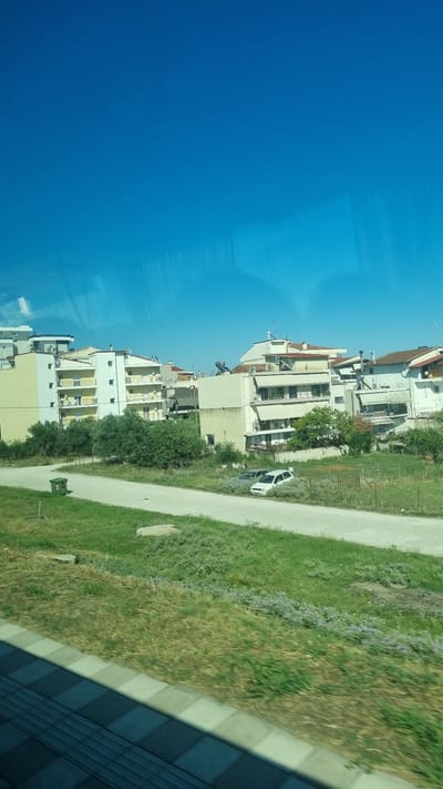 The image captures a view of residential buildings under a clear blue sky, likely from a moving vehicle given the slight blur in the foreground. The buildings are multi-story, painted in light colors such as yellow and white, with balconies. Greenery, including trees and grass, surrounds the buildings. A dirt road runs in front of the buildings, with two cars parked on it and a green trash bin to the left. The scene appears to be in a suburban or semi-urban area. The presence of the specific architectural style of the buildings and the characteristic Mediterranean vegetation suggest a location in Greece. Specifically, the image is consistent with a scene in the city of Larissa, Greece, which has similar residential areas and landscapes.