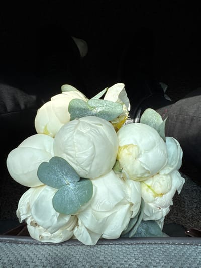 A bouquet of white peonies and eucalyptus leaves rests on a textured surface, possibly a car seat or fabric-covered furniture. The flowers are tightly clustered, with some buds partially open and others still closed. The eucalyptus leaves are a muted green and appear to be interspersed throughout the bouquet. The background is dark and out of focus, suggesting the photograph was taken indoors or in low light conditions, possibly within a vehicle. The lighting casts shadows on the flowers, highlighting their delicate texture and form. There is no identifiable urban or natural landscape visible in the image, making it impossible to pinpoint a location within the city of Imeni Chapaeva, Kyrgyzstan. The scene is static and depicts a still life arrangement of flowers.