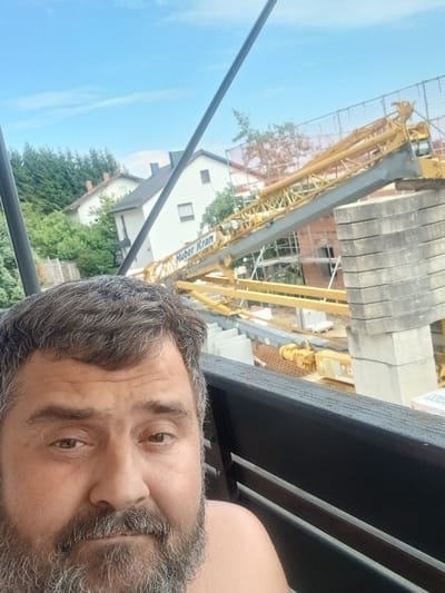The media file is a close-up, portrait-oriented photograph of a man's face, taken from a slightly high angle, with a construction site visible in the background. The man has a beard with graying hairs and is looking directly at the camera with a neutral expression. His eyes are open, and his gaze is steady. The lighting suggests it is daytime, likely morning or afternoon, with natural light illuminating his face.
In the background, a large yellow construction crane dominates the scene, its lattice structure extending upwards. The crane has the text "Huber Kran" visible on its boom. Construction materials, including concrete blocks, are stacked near the base of the crane. Behind the construction site, residential buildings with white facades and tiled roofs are visible, interspersed with lush green trees. The sky is a clear, bright blue, indicating fair weather. The overall setting appears to be a residential area undergoing construction. The orientation of the photograph is such that the man's head is on the left side and the background elements are on the right.
