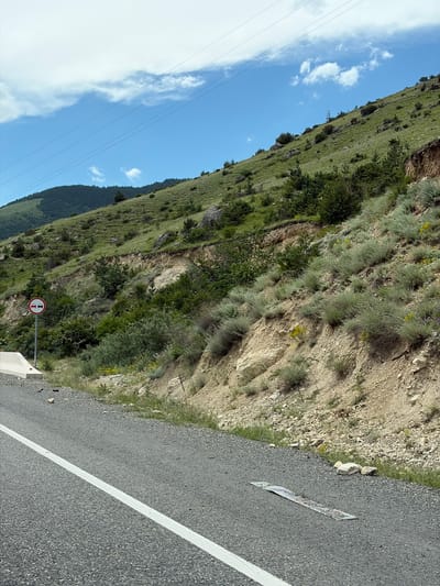 The image shows a roadside view on a steep, vegetated hillside. The road is paved asphalt with a white lane marking. A white concrete barrier is visible on the left side of the road. A circular traffic sign with a red border, a white background, and two red car symbols with a red bar across them is mounted on a pole to the left of the road. The hillside is covered with green grass, shrubs, and scattered trees, with exposed earth and rocks in some areas. Power lines are visible stretching across the sky. In the distance, a mountain range is partially obscured by the hillside. The sky is blue with scattered white clouds. The scene is consistent with a rural or mountainous area. The mention of Zintsar, Russia in the prompt appears to be contextual information and does not directly provide visual cues within the image itself. There are no people or visible actions taking place.