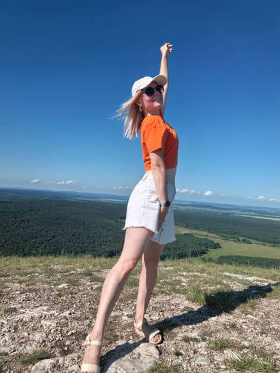 A young woman with blonde hair stands on a rocky, grassy hill under a clear blue sky. She is wearing an orange crop top, a white denim skirt with a frayed hem, a white baseball cap, and sunglasses. Her right arm is raised, and she is looking to her left with a smile. Her left hand is placed on her skirt. She is wearing tan sandals. In the background, a vast landscape of green forests and rolling hills stretches out to the horizon. The location is identified as Shikhan, Russia.