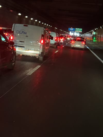 This media file captures a traffic jam on a highway in Noisy-le-Sec, France. The scene is set at night, possibly in rainy weather, as evidenced by the reflections on the wet road and the illuminated streaks appearing as water droplets. The primary subjects are a line of vehicles, predominantly vans and cars, stuck in congestion within a tunnel. The vehicles' brake lights are illuminated, indicating a standstill or slow-moving traffic.
Notable details include several traffic signs visible on the left side of the tunnel, suggesting it is an urban or suburban highway. One sign prominently displays green rectangular shapes, possibly indicating lane guidance or exit information. Another sign has blue text, reading "NOISY LE SEC CENTRE," confirming the location.
The overall atmosphere is one of typical rush-hour or post-event congestion, with no obvious signs of distress or emergency, just the mundane reality of traffic delays. The lighting within the tunnel, provided by overhead fixtures and vehicle lights, creates a dark and somewhat enclosed environment.
Text visible on one of the vans includes "OLFM" in large blue lettering, and a license plate reading "0-083-PME". Another visible phone number is "06 99 46 93 10". The time of day is clearly night due to the darkness and artificial lighting. The reflections and streaks on the road suggest precipitation, adding a slightly moody feel to the scene.