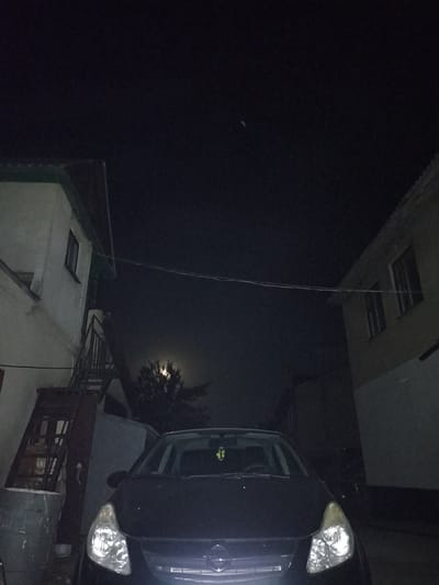 The image depicts a dark night scene in a narrow alleyway or courtyard, possibly in the city of Pochaiv, Ukraine. On the left, a light-colored building with a green-trimmed roof is visible, featuring a window and an exterior staircase. On the right, a beige building with dark windows is seen. A dark-colored car, possibly a hatchback, is parked in the center of the frame, its headlights illuminated. In the background, a tree with foliage is partially obscuring a bright full moon. The overall atmosphere is dimly lit and appears to be a residential or service area.