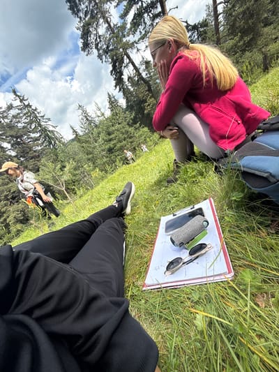 Two individuals are on a grassy hillside in a wooded area, likely in Yundola, Bulgaria, based on the context provided. In the foreground, a person is lying down, their legs clad in black pants, with a notepad, a smartphone, a small cylindrical object, and sunglasses placed on the grass. In the background, a woman in a pink jacket and light-colored leggings is crouched, looking towards the left. Further up the hill, two other individuals, one wearing a light-colored hat and white shirt, are visible in the distance. The environment consists of lush green grass and numerous evergreen trees, with a cloudy sky overhead.