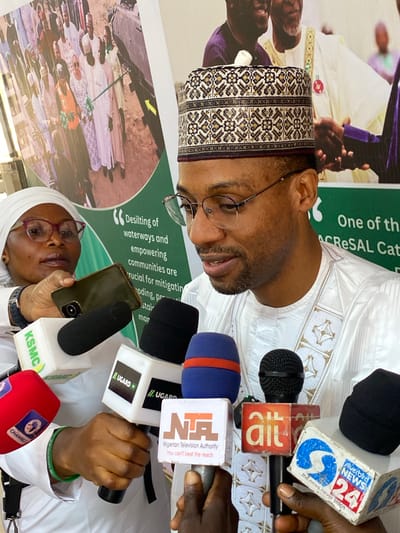 This image captures a media briefing or press conference held in Kwarau, Nigeria. The central figure is a man, likely a dignitary or official, wearing traditional Nigerian attire consisting of a white embroidered agbada and a patterned cap. He also wears glasses and has a neatly trimmed beard. He is seen speaking into a cluster of microphones, indicating an official statement or address to the press.
Surrounding the speaker are numerous microphones held by various individuals, representing different media organizations. Visible logos on the microphone covers include "silverbird NEWS 24" with the slogan "the news never stops," "ait," "NTA Nigerian Television Authority" with the slogan "You can't beat the reach," "UGARD TV," "KSMC," and "CHANNELS." In the foreground, a woman wearing a white hijab and glasses is partially visible, holding a smartphone, seemingly recording the event. One hand of a person holding a microphone wears a green wristband with "UGARDW" visible.
Behind the speaker, a large banner is displayed, featuring text that reads: "Desilting of waterways and empowering communities are crucial for mitigatin oding, pr stair mar" and "One of th ACRESAL Cat." This suggests the event is related to environmental or community development projects. Another section of the banner shows a large circular image of several people, possibly indicating community engagement or project beneficiaries. The lighting suggests it is daytime. The overall scene conveys a formal and informative gathering, characteristic of an official announcement or public address.