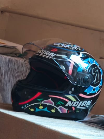 A black Nolan motorcycle helmet with a clear visor is positioned on a textured surface, possibly a piece of furniture. The helmet is adorned with colorful graphics, including blue floral patterns, a prominent white number "7" within a blue circle, and a red stripe. The "Nolan" logo is visible on the side in white lettering. The background consists of a textured wall and what appears to be stacked papers. The overall lighting suggests an indoor setting with natural light. This image is not located in Tsarev Brod, Bulgaria; it depicts a helmet.