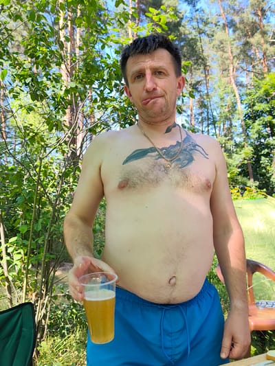 In the city of Horods'ke, Ukraine, a shirtless man with a tattoo of a scorpion on his chest and a gold chain around his neck stands outdoors. He is holding a plastic cup filled with a light-colored beverage, possibly beer, in his right hand. The man is wearing blue swim shorts. His facial expression is one of amusement, with his tongue slightly extended. The background consists of trees and foliage, indicating a natural or park-like setting. A bright green tent and an orange plastic chair are partially visible to the right. The overall scene suggests a casual outdoor gathering or recreation.