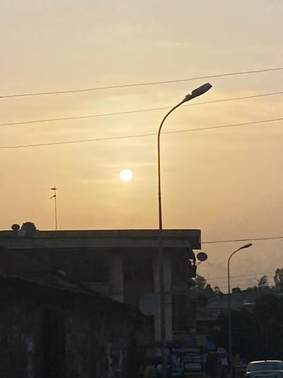 The image shows a landscape in Abofifia, Nigeria, at sunset. The sun is visible in the center of the frame, with the sky appearing in shades of orange and yellow. A street light is positioned to the left of the sun, with power lines visible in the sky. Buildings and trees are seen on the right side of the image. There are no people visible.