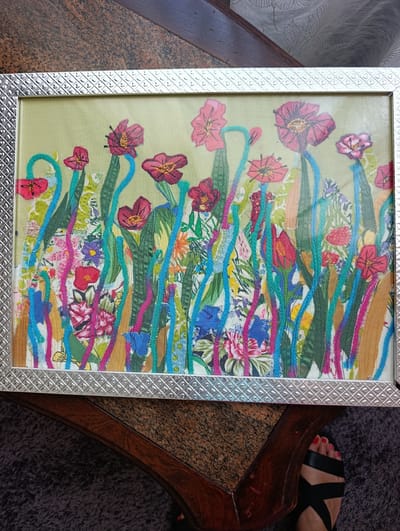 This media file shows a framed piece of fabric art depicting a vibrant, abstract floral scene. The artwork is a collage of colorful fabrics and embroidery, featuring stylized flowers and foliage in shades of red, pink, green, blue, yellow, and white. The floral elements are layered and intertwined, creating a dense and energetic composition. Some of the flowers appear to be poppies, with their characteristic cup-shaped petals.
The artwork is encased in a silver-toned frame with a textured, geometric pattern along its inner edge. The background of the artwork is a pale, textured fabric, possibly linen or a similar material, in a light green hue. The fabric elements and embroidery create a tactile and visually rich surface.
The framed artwork is placed on a table with a dark wood and speckled brown surface. The table appears to be outdoors or near a window, as evidenced by the lighting and the presence of a person's feet in the lower right corner of the image. The person is wearing black sandals and has red-painted toenails, suggesting a warm or pleasant weather condition. The overall impression of the artwork is one of joyful exuberance and natural beauty, rendered in a bold and modern artistic style. There is no visible text in the image. The location context of Odivelas, Portugal, does not provide specific clues within the image itself.