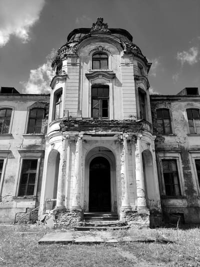 The image depicts a decaying, multi-story building with classical architectural elements. The structure is severely dilapidated, with crumbling walls, broken windows, and weathered surfaces. The exterior features arched doorways, ornate detailing, and decorative trim. The facade shows significant signs of disrepair, including exposed brick and missing sections of the original structure. The surrounding environment includes grassy areas and a sky with scattered clouds. The image appears to be taken in Жалудоцкі сельскі Савет, Belarus.