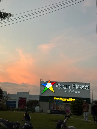 An exterior shot of the Gran Plaza San Antonio shopping center in Pitalito, Colombia, is captured during dusk. The sky transitions from a light blue-grey at the zenith to pronounced orange and pink hues at the horizon, indicating sunset conditions. Sparse clouds are present. In the upper left quadrant, dark fronds of a palm tree are partially visible along with three parallel overhead power lines. The main building features a large, illuminated sign reading "Gran Plaza San Antonio" in white text, accompanied by a multi-colored star logo (white, blue, green, red, orange, yellow sections) to its left. Below the main sign, a secondary illuminated sign displays "Dollarcity" in yellow lettering. The building's facade consists of white and dark green sections, with a large blue-tinted window and a red banner visible on the left side. The foreground presents a sloping grassy area. Partially visible on the left foreground are two dark-colored motorcycles or scooters. On the right, three individuals are present: one male in a brown long-sleeved shirt and light-colored pants is observed holding a mobile phone to his ear, facing right. Another person in dark clothing is partially visible further right, and a smaller, darker figure is near the building's base.