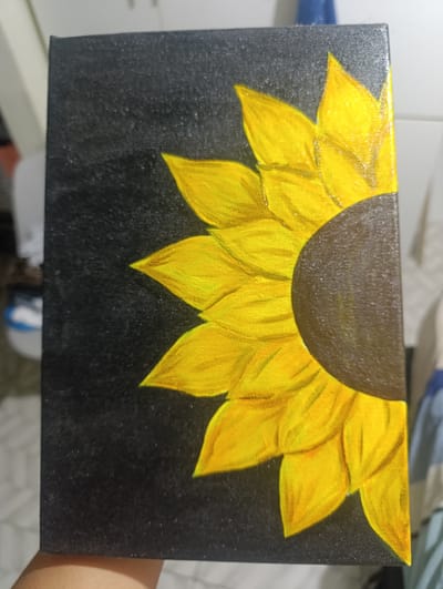 A close-up photograph shows a painting of a half-sunflower on a canvas. The sunflower is painted with shades of yellow and brown, contrasting with a black background. The canvas's texture is visible, and it is held by a hand. The location is unknown and not discernible from the image.