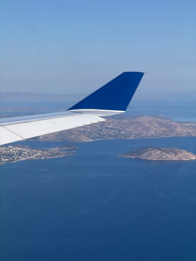 The image shows an aerial view from an airplane window. Part of the plane's tail fin, colored blue and white, is visible in the left foreground. Below, there is a body of water with land formations, likely islands or peninsulas, suggesting a coastal area. The land shows signs of vegetation and possible human settlement. The sky is clear and blue. Based on the location, it's probable that the picture was taken over the Paiania Municipal Unit, Greece, given the presence of islands in the sea.