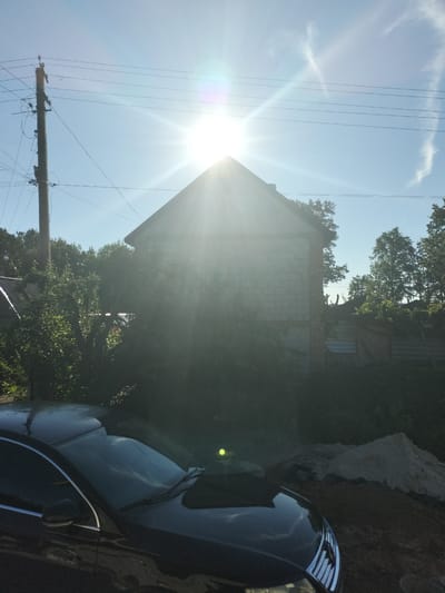 The image depicts a sun-drenched exterior scene, likely in Kudashivka, Ukraine, on a clear day. A prominent, bright sun is positioned high in the sky, causing lens flare and obscuring parts of the scene with its intense light. In the foreground, the hood and part of the roof of a dark-colored car are visible, reflecting the sky and surroundings. Behind the car, a building constructed from light-colored cinder blocks with a gabled roof stands partially shrouded in foliage. Electrical wires run across the sky, supported by a wooden utility pole to the left of the frame. Dense green trees and bushes surround the building and extend towards the edges of the image. To the right of the building, parts of a wooden fence and what appears to be a pile of sand or dirt are visible. The overall impression is one of a rural or semi-rural setting under bright daylight.