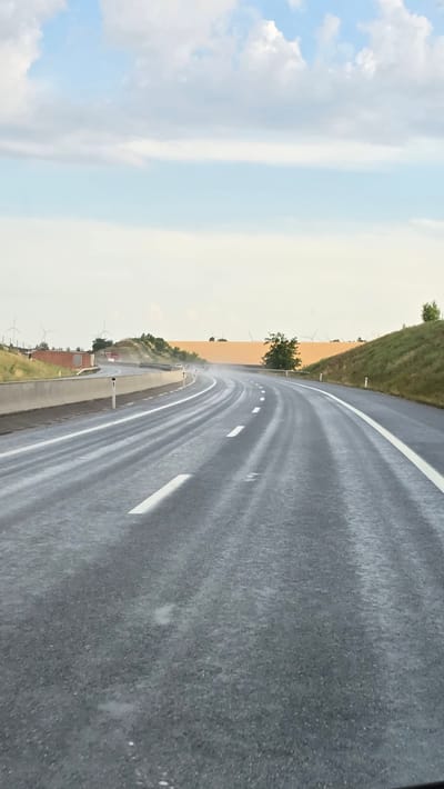 The image shows a wet highway in Hobersdorf, Austria, with visible rain spray and puddles on the asphalt. The road curves to the right, with white lane markings. On the left side of the road, there is a concrete barrier, a grassy embankment, and a few wind turbines in the distance. On the right side of the road, there is a grassy hill. The sky is cloudy with patches of blue. The overall impression is one of a rainy, overcast day on an open highway.