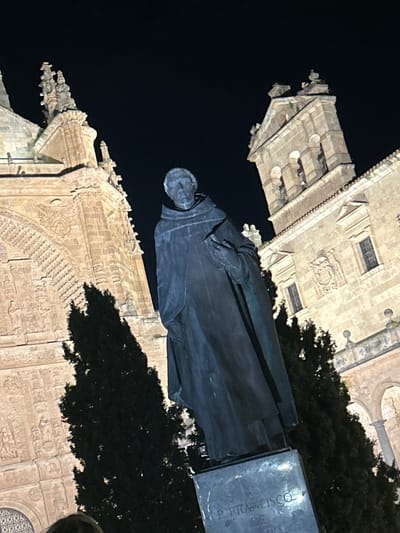 The image depicts a nighttime scene in Salamanca, Spain, specifically featuring the statue of Fray Francisco de Vitoria. The central subject is a weathered bronze statue of a male figure dressed in monastic robes, holding a book in his left arm. The statue stands on a dark stone pedestal, where the inscription "P. FRANCISCO DE [VITORIA]" is partially visible.

In the background to the left, a section of the illuminated Salamanca Cathedral complex is visible, characterized by highly ornate Plateresque architectural details, intricate carvings, arches, and a series of pinnacles. To the right, another illuminated historical stone building, likely part of the University of Salamanca or an adjacent structure, features arched windows, a coat of arms embedded in its facade, and a prominent bell tower with arched openings. Several tall, dark green coniferous trees are interspersed throughout the scene, positioned behind the statue and partially obscuring the lower sections of the background buildings. The sky is completely dark, indicating night, and the scene is brightly lit by artificial floodlights, emphasizing the architectural details and the statue. The perspective is low-angle, looking upwards at the statue and the structures.