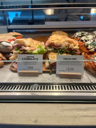 This image captures a close-up view of a refrigerated display case in a food establishment, likely a deli, cafe, or bakery, in Dorno, Italy. The case holds several freshly prepared food items, primarily focaccia or pizza alla pala sandwiches.
In the foreground and midground, two distinct sandwich types are prominently displayed, each with an accompanying white price tag and wooden stand. The top sandwich, labeled "TRICOLORE," features prosciutto di Parma, fiordilatte mozzarella, cherry tomatoes, and rocket salad. The sandwich below it, labeled "COTTO E FIORDILATTE," contains cooked ham, fiordilatte mozzarella, and lettuce. Both items are priced at € 8,20. The descriptions on the tags are in Italian and English, detailing the ingredients of each "Pizza alla pala" (paddle pizza or flatbread).
Behind these items, a large, rectangular pizza or focaccia with a reddish topping (possibly tomato sauce and other ingredients) is visible. The display case itself is made of glass and stainless steel, with reflections visible on the left side, including what appears to be part of the word "CATALI" and a blurred reflection of a person. The setting suggests a casual dining or takeaway environment where fresh Italian specialties are offered.
