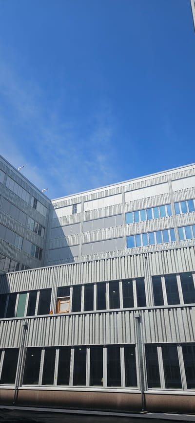 The image shows a modern, multi-story building with a facade of metallic louvers and numerous windows. The building appears to be an institutional or commercial structure, possibly an office building or a school, characterized by its clean lines and repetitive design elements. The windows are a mix of clear and tinted glass, with some showing reflections of the bright blue sky. Several windows on the upper floors have a reflective blue tint. On a lower level, there are larger, darker windows that appear to be opaque or heavily tinted. The building's exterior is predominantly silver or light gray, suggesting a contemporary architectural style.
The scene is set against a clear, bright blue sky with a few wispy white clouds. The angle of the photograph suggests it was taken from ground level, looking upwards at the building. The lighting indicates it is daytime, likely with the sun casting shadows on the building's facade, highlighting the texture of the louvers.
This building is located at the Kronan area in Västerås, Sweden, specifically the Cronqvist building of Mälardalens University. The Cronqvist building is part of the university's campus expansion and is designed to accommodate various academic departments and student facilities. The metallic exterior and numerous windows are characteristic of modern educational institutions aiming for a bright and open learning environment. The construction or renovation aspects hinted at by the exposed window frame on the second floor may indicate ongoing work or maintenance at the site.