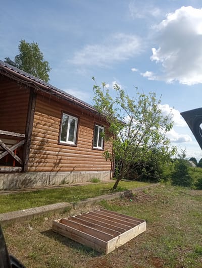 This media file shows a wooden house with a porch, situated in a grassy area with trees and a bright blue sky filled with white clouds. A wooden structure, possibly a frame or a planter box, is visible on the right side of the frame. The house is constructed from logs and has two white-framed windows. A tree with green leaves is growing near the house. The scene appears to be outdoors, possibly in a rural setting in Belarus, given the location context. The lighting suggests it is daytime, and the weather seems pleasant with sunshine and clouds. There is no visible text in the image.