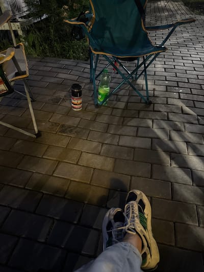 The image depicts a ground-level view of a paved outdoor area at night. Two folding chairs, one predominantly teal with orange trim and the other partially visible and light-colored, are present. A bottle of green liquid, possibly a beverage, sits near the teal chair. A cylindrical container, black with colorful labels, stands upright on the paving stones. The ground is covered with dark, rectangular paving stones, showing some wet patches and casting long shadows from an unseen light source. In the foreground, a person's legs and feet are visible, wearing light-colored pants and athletic shoes with a combination of white, dark blue, and green accents. The background shows dark foliage and possibly a large, industrial-looking fan or ventilation unit. The overall impression is of a casual, outdoor setting, potentially a campsite or a park area.