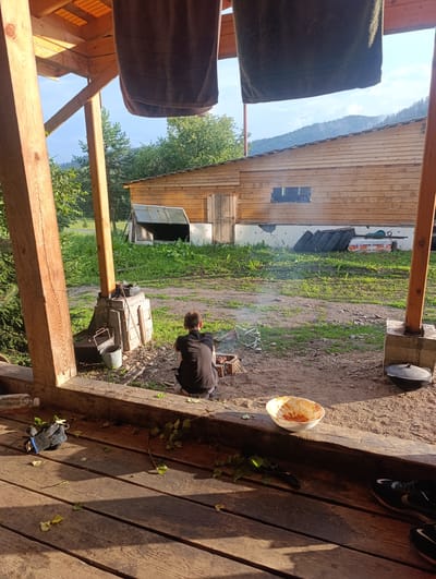 The image depicts a rustic outdoor scene, likely at a dacha or rural residence in Biryuchul, Russia. In the foreground, a wooden porch is visible, with scattered leaves and a pair of black athletic shoes. A white bowl containing remnants of food sits on a wooden beam. In the middle ground, a person is crouched down near a small barbecue, tending to a fire. Various items are scattered around the barbecue area, including a bucket and grill grates. A rustic wooden building with a large opening is situated in the background. The area surrounding the building is grassy and dirt-covered. The scene is bathed in the warm light of what appears to be late afternoon or early evening sun. The overall impression is one of a casual outdoor gathering or meal preparation in a natural, somewhat unkempt setting.