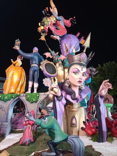 An elaborate, multi-tiered artistic installation, likely a Fallas monument, is depicted at night in El Port De Sagunt, Spain.
In the foreground, a large, stylized female bust figure resembling a queen or witch, with pale skin, exaggerated eyes, a purple crown, and a purple gown, dominates the right side. She holds a purple handheld mirror with a reflected smiling face in her right hand, and a brown, donut-shaped object with green dripping liquid (resembling a poisoned apple or confection) in her left. Below and to her left, a wolf-like character in a green suit and brown fedora is bent forward. Behind him, a smaller female figure in a long pink dress stands, facing left.
Above these figures, on a grey castle-like structure featuring archways and green foliage details, a male figure in a blue suit is seen from behind, holding a golden lantern or bell jar containing a red rose aloft in his right hand. To his left, a female figure in a voluminous yellow dress is partially visible. Integrated into the mid-section of the structure are two small, dark, bat-like creatures with red wings.
At the top of the monument, a dynamic figure in striped clothing, possibly riding a broomstick, is depicted flying upwards, surrounded by various fruits and objects attached to the broom. The entire monument is brightly illuminated by artificial lights against a dark, featureless night sky. The base of the monument is surrounded by white aggregate material and artificial green foliage.