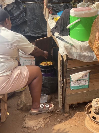 The image captures a vibrant outdoor scene, likely in Ojodu, Nigeria, featuring a person engaged in cooking during the daytime. The perspective is from above, looking down on the individual and their surroundings.

The main subject is a person, likely a woman, visible from the upper thigh to the top of her head. She wears a light-colored (possibly white or off-white) t-shirt and light-colored shorts or skirt. Her right arm is extended over a cooking pot, where she is stirring a dish that appears to be fried plantains or bananas, glowing golden in the pan. The heat source is not fully visible but is beneath the pot. On her feet, she wears brown sandals with distinct white buckles.

The setting is an informal outdoor cooking area. The ground is made of reddish-brown dirt, with scattered small stones and debris. Various items are placed on wooden planks that serve as makeshift tables or surfaces. To the left, a large green and white cooler (ice chest) sits atop a black plastic sheet and white lace fabric. A kitchen knife rests nearby on the white fabric. Further right, a rustic wooden surface holds a green document or paper. Part of a rusty metal wheel rim is visible in the upper right corner, resting on the dirt ground.

The green document prominently displays text, including "QPoy Beyond Banking..." and "Developed by... for OPay...". A sequence of numbers is partially visible, including "65481217" and "217", possibly a phone number or account identifier. The natural light indicates it is daytime, and the distinct shadows suggest a sunny day. The overall scene depicts a common daily activity in a simple, functional environment.