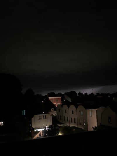 The image captures a dark, ominous sky over a city at night, with a thunderstorm visible in the distance. The foreground is dominated by the silhouettes of buildings and trees. A large, distinct building with multiple windows is visible on the left side, with a red-colored building with white-trimmed windows to its right. In the center and right of the image, a row of white buildings with arched roofs and illuminated windows are partially visible, suggesting some activity within. The overall atmosphere is one of a brewing storm in a residential or urban area. This scene is consistent with the view from a residential area in Deventer, Netherlands, particularly during a stormy night.