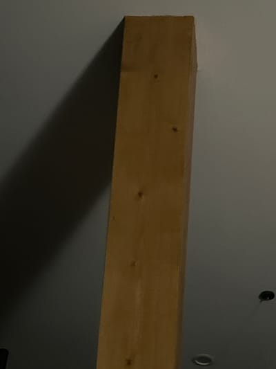 The image shows a close-up of a light-colored wooden beam positioned vertically in the center of the frame, casting a shadow on a grey wall to its left. The beam has visible knot details. The background is a plain, lightly textured grey surface, possibly a ceiling or wall. There are no people or discernible activities in the image. The scene appears to be an interior space, with no specific urban context of Siedlce, Poland, identifiable.