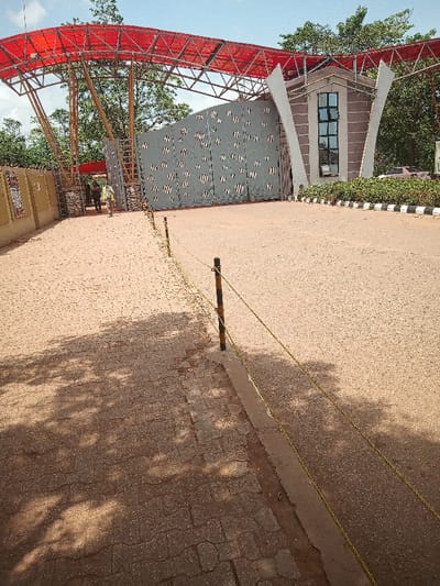 The image shows an empty, paved driveway leading to a closed gate, situated in front of a building at the coordinates 6.682, 3.168 in Otta, Nigeria. The gate is made of grey metal and has a white and black striped pattern. A red, metal canopy is installed over the entrance of the driveway. The gate is closed and appears to be secured, with no visible access point. There are three individuals walking toward the gate, but they have stopped and seem to be waiting. The driveway is bordered by a black and white curb and a grassy area with small trees and bushes.  A line of yellow rope, secured to black posts, runs parallel to the sidewalk. The sun casts shadows on the pavement, and the sky is clear and blue. The scene is devoid of traffic, but the presence of the individuals and the gated entrance suggest the possibility of an active, functional facility within the premises.