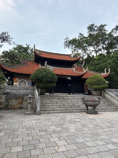 This media file shows the exterior of a temple in Hy Cuong, Vietnam. The temple features traditional architecture with a red-tiled, curved roof adorned with decorative elements. A stone staircase leads up to the temple entrance, which has dark wooden doors. To the right of the staircase, a large, ornate incense burner sits on a stone pedestal.

The surrounding area is lush with greenery, including mature trees and well-maintained bushes, suggesting a temperate or tropical climate. The sky is overcast, indicating a cloudy day. The architecture and the presence of the incense burner point to a religious or cultural site, likely a Buddhist temple given the Vietnamese context.

Notable details include the intricate carvings on the stone structures and the overall serene atmosphere of the location. The image is oriented vertically, giving a unique perspective on the temple and its surroundings. No people are visible in the frame.

Visible text includes signage on the temple building, with some characters appearing to be Vietnamese. One sign reads "SẮP LỄ" which translates to "Ceremony About To Begin" or "Upcoming Ceremony".