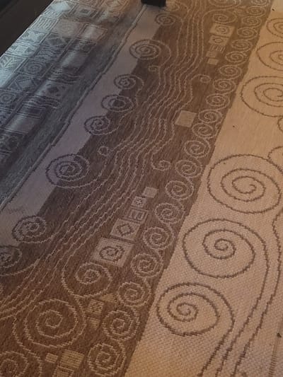 The scene presents an angled, close-up view of a woven textile, consistent with a floor rug or mat. The textile features multiple vertical bands of contrasting patterns and tones in a palette of browns, beiges, and creams. The leftmost visible band exhibits geometric square and X-shaped motifs. Adjacent to it, a wider band displays a complex arrangement of wavy lines, smaller square elements, and spirals. To the right, a prominent band showcases larger, distinct spiral patterns against a lighter background. The weaving appears to be a coarse, flat-weave texture. Uneven lighting is present, casting a slight shadow over the upper left portion of the textile. No individuals, additional objects, specific actions, or interactions are visible. The surrounding environment is not discernible beyond the textile itself. The image does not contain data to specify the location within Κωστήτσι, Greece.