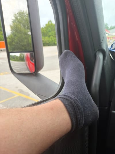 A person's leg with a gray sock is resting on a red interior surface inside a vehicle. The person's ankle and lower leg are visible, with light hair on the skin. The exterior of the vehicle is partially visible through the window, showing a side mirror reflecting trees and a red truck. Outside, yellow lines mark parking spaces on an asphalt surface, and a blurry building with red and yellow accents is in the background. The scene appears to be a parking lot in Wildwood, United States.