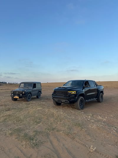 Two black vehicles are parked on a dirt road. The vehicles are facing the camera. The vehicle on the left is a dark gray Mercedes-Benz G-Wagon. The vehicle on the right is a black Ram 1500 TRX. The vehicles are parked in an open field with a blue sky and some clouds in the background. The field is mostly dirt, but there is some vegetation visible. The location of the image is Bnei Zion, Israel at latitude 32.226, longitude 34.849. The vehicles are both facing the camera. The Mercedes-Benz G-Wagon is slightly angled to the left. The Ram 1500 TRX is angled slightly to the right. The front of the Mercedes-Benz G-Wagon has a license plate with the number 190-95-65 on it. The Ram 1500 TRX has the RAM logo on its front grille. Both vehicles are in good condition.