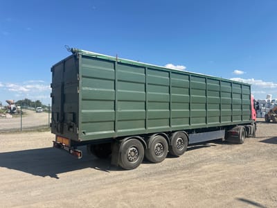 This media file shows a green, flatbed truck with a large, open-top cargo container. The truck is parked on a gravel lot under a clear blue sky with a few wispy clouds. The sun appears to be high in the sky, indicating it is likely midday.
The cargo container has a green tarp covering its top, held down with ropes and metal clasps. The sides of the container are made of green metal panels with visible horizontal ridging. The truck has a red cab and a long wheelbase with multiple sets of dual tires. The Scania logo is visible on the rear mudflap. The rear of the truck features a yellow and red striped bumper with integrated lights.
In the background, a wire fence encloses an area with some industrial equipment and a white van. Further in the distance, more industrial elements and a yellow construction vehicle are visible. Large white sacks are piled up to the right of the truck.
The scene appears to be an industrial or construction area, possibly a storage yard or a loading/unloading zone. The overall impression is one of a functional, utilitarian setting, with the truck ready for transport or having recently completed a delivery. There is no visible text on the truck or the cargo container, other than the Scania logo.