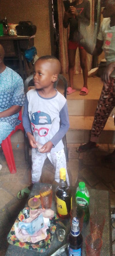 The image depicts a casual indoor gathering, likely in a domestic setting in Ohafia, Nigeria. In the foreground, a young Black boy with a buzz cut stands, looking to his left with a neutral expression. He wears a light grey and blue long-sleeved shirt featuring a design with a cap bearing the words "COOL" and "COLLEGE," and large red-framed glasses. His pants are light-colored with subtle patterns. He holds a small, light-colored item, possibly money, in his hands.
To the boy's left, an adult person is partially visible, seated on a red plastic chair and wearing a blue and white patterned top. In the background, on raised concrete steps, another person (likely female) stands, visible from the waist down, dressed in a dark top, red shorts, and pink slide sandals. This individual appears to be holding a mobile phone. Another person's legs, wearing leopard print pants, are partially visible on the right.
A table in the immediate foreground displays several items: a dark liquor bottle with a prominent yellow label showing "STAR," a green plastic bottle with "LIMAX" partially visible (likely a soft drink or mixer), and a beer bottle with a blue and white label. Several empty glasses are also present. A decorative plate holds crumpled Nigerian Naira notes, including a blue 200 Naira bill, alongside some small snacks or offerings. In the deeper background, on another table, a black electronic device, possibly a radio or speaker, is visible. The lighting suggests it is daytime. The overall atmosphere is informal and relaxed.