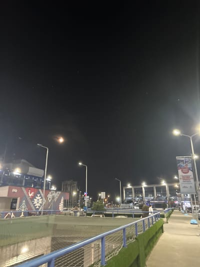 The image captures an urban night scene, likely in Bayrampaşa, Turkey, characterized by artificial illumination against a dark sky. A prominent bright light source, possibly the moon or a strong distant light, is visible in the upper-left sky, surrounded by numerous smaller, scattered bright points that appear to be distant lights or stars.
In the foreground, a blue metal railing or fence with a mesh pattern runs diagonally, separating a paved area from a strip of green artificial grass. Beyond this, several buildings with illuminated windows are visible. One building particularly stands out with a large, colorful mural on its side, featuring a geometric pattern reminiscent of traditional Turkish carpets (Kilim) in shades of red, white, and blue.
Numerous streetlights, equipped with bright lamps, line the street, casting strong glows and illuminating the surroundings. A large banner hangs from one of these streetlights. The visible text on the banner is in Turkish and reads: "Şimdi öğrencilere 2.500 TL burs desteği veriyoruz!" (Now we give 2,500 TL scholarship support to students!) and "DAHA İYİ GELECEK İÇİN BİRLİKTE YÜRÜYORUZ" (We walk together for a better future). The word "BÜYÜKŞEHİR" (Metropolitan Municipality) is also discernible, indicating the message likely comes from a municipal body. Another building has a sign that partially reads "DAN-SMA / information".
A single person is partially visible in the midground, standing near the fence, wearing a light-colored shirt or jacket. The time of day is clearly night, with the artificial lights providing the primary illumination. The scene depicts a quiet urban environment with commercial or public buildings.