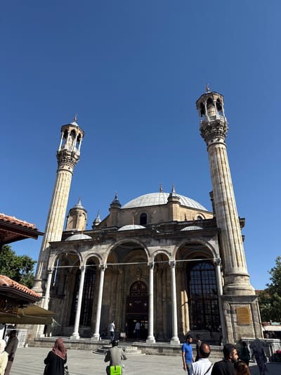 This is a low-angle, exterior, eye-level, full shot of the Aziziye Mosque in Karatay, Turkey, captured on a clear, bright day. The mosque features a central dome and is flanked by two tall, ornate minarets. The building's facade is characterized by a series of arched walkways supported by columns. People are visible in the foreground and near the entrance of the mosque, with some walking, others standing, and one person riding a scooter. The environment appears to be an open plaza or courtyard in front of the mosque, with glimpses of trees and parts of other structures to the sides, including a tiled roof on the left. The sky is a solid, deep blue.