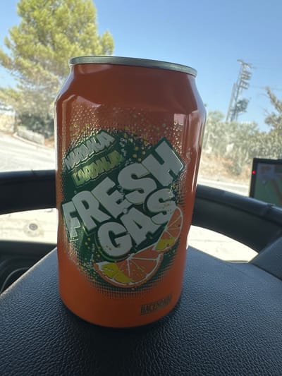 An orange soda can, labeled "NARANJA LORANHA FRESH GAS," sits on a black, textured surface, likely a car's dashboard. The can features green graphics with orange slices and white lettering. In the background, a bright green tree is visible against a clear blue sky, with some power lines and a blurry vehicle interior suggesting an outdoor, possibly roadside setting. The label "HACENDADO" is visible on the lower portion of the can. The scene is well-lit, indicating daytime.