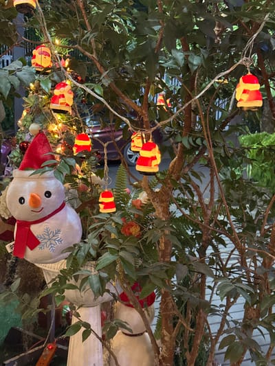 The scene depicts an outdoor nighttime view of Christmas decorations interspersed among natural foliage in Buôn Ma Thuột, Vietnam. Multiple strands of illuminated string lights, featuring plastic Santa Claus head figures and generic red and yellow stacked shapes, are strung through the branches of several trees and bushes with green leaves. In the lower-left foreground, a white plush snowman, wearing a red pointed hat, an orange nose, black dot eyes, and a red scarf with a light blue snowflake emblem, is situated on a white fluted pedestal. Part of a second plush snowman, similarly adorned, is partially visible behind the foreground elements. Behind the decorated foliage, a dark-colored vehicle, likely a car, is partially visible, with its front wheel and part of its body discernible on what appears to be a street or driveway. The background also shows indistinct structural elements and additional green plants.