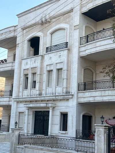 This image presents a detailed, sideways view of the facade of a multi-story building in Aleppo, Syria, captured under bright daylight. The building showcases a classical architectural style, with light-colored, possibly stucco or stone, exterior walls.
The facade is rich in detail, featuring numerous arched openings. Many of these are fitted with closed, ridged roller shutters, indicating windows or balcony access. Several small balconies, some recessed and others slightly projecting, are adorned with ornate black wrought-iron railings displaying geometric and scroll patterns. Rectangular windows with dark frames are also visible across different levels, some appearing as single units and others as pairs.
A prominent feature on a lower level is a large, dark, and intricately designed entrance door, framed by classical architectural elements. The building incorporates decorative pilasters, cornices, and raised rectangular panels, contributing to its grand aesthetic. At least one traditional-style wall lamp with a dark fixture and a bell-shaped shade is mounted on the facade, adding to its classic charm.
Several black electrical wires run across the building's exterior, indicating utility connections. Sparse green foliage, likely from tree branches, is visible at the top edge of the image. The sky is clear and light blue, suggesting fair weather during the day, likely mid-morning or afternoon given the soft lighting. The building shows some signs of weathering and discoloration on ledges and under balconies, yet its intricate details and overall structure remain well-preserved. There are no people or discernible text in the image.