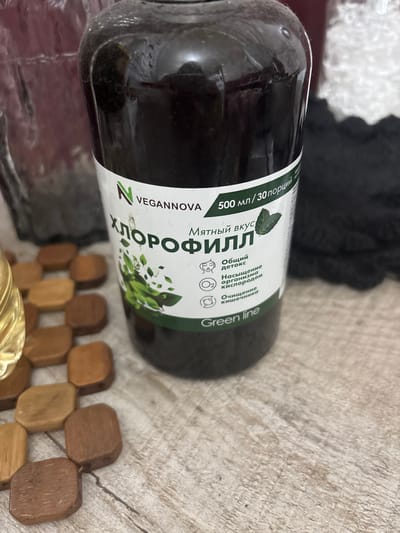 A dark glass bottle of liquid is placed on a wooden surface. The bottle has a white label with green text and graphics. The text is in Russian and reads "ХЛОРОФИЛЛ" (Chlorophyll) and "500 мл/30 порций" (500 ml/30 servings). Below this, it lists benefits such as "Общий детокс" (General detox), "Насыщение организма кислородом" (Oxygenation of the body), and "Очищение кишечника" (Intestinal cleansing). The brand name "VEGANNOVA" is visible at the top of the label. A brand logo with a green "N" is also present. The bottom of the label reads "Green line". To the left of the bottle are several light brown wooden blocks arranged in a grid pattern. The background is blurred but appears to be a continuation of the wooden surface and a dark fabric-like material. There is no visible indication of the city of Княгинино, Russia.