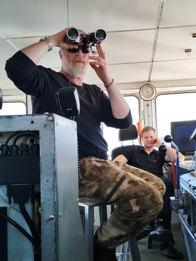 Two men are on the bridge of a ship. The first man stands, looking through binoculars. He wears sunglasses, a black long-sleeved shirt, and camouflage pants. A watch is on his left wrist, and a bracelet on his right. He has a full white beard. The second man sits in a chair. He wears a black shirt and black pants, and black clog-style shoes. His left hand rests on his chin. A clock is visible on the bridge. The location is unknown.