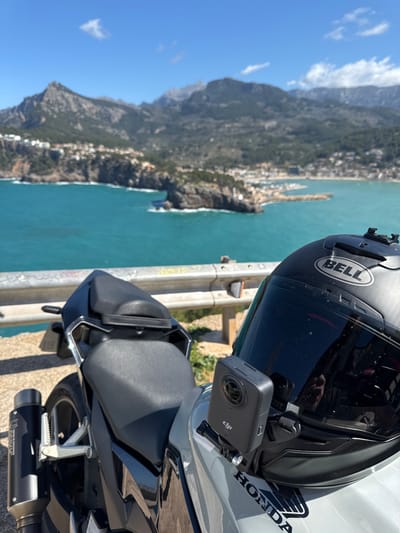 A motorcycle helmet, branded "BELL" and featuring a dark tinted visor, rests on the tank of a light-colored "HONDA" motorcycle in the foreground. A black DJI action camera is mounted to the right side of the helmet. Behind the motorcycle and helmet, a metal guardrail, showing some wear and markings, runs horizontally. Beyond the guardrail, a panoramic view of Port de Sóller, Spain, unfolds under a clear blue sky. The turquoise waters of the Mediterranean Sea dominate the midground, with a prominent rocky headland featuring a natural arch formation extending from the left into the bay. Waves are observed breaking against this natural formation. To the right, the town of Port de Sóller is visible along the coastline, with a marina containing boats and buildings lining the shore. In the background, the rugged, vegetated peaks of the Serra de Tramuntana mountains rise behind the coastal settlement. The scene is brightly illuminated by natural sunlight, indicating clear weather. No individuals are present in the immediate vicinity.