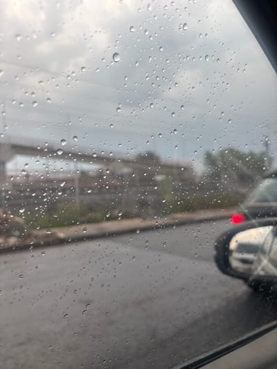 The image shows a view through a rain-covered car window. The raindrops obscure the scene outside, but a red light is visible in the reflection of a car side mirror. In the background, a blurred building or structure is visible, along with power lines. The overall impression is of a rainy day in an urban setting. There are no people visible. The city of Tultitlán, Mexico cannot be determined from this image.