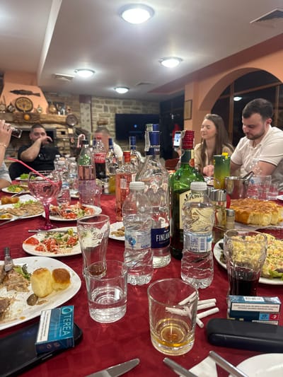 An indoor scene in a restaurant or tavern in Arkovna, Bulgaria, depicts a social gathering around a rectangular table draped with a red tablecloth. Overhead circular lights illuminate the space. The background features a stone wall, a large decorative fireplace structure adorned with various objects including clocks and pottery, and an arched doorway leading to another area. A flat-screen television is mounted on a distant wall.

On the table, numerous plates with partially consumed meals are present, including grilled meat, small bread rolls, and several salads (one appearing to be a mayonnaise-based salad, another a tomato and cucumber salad). A large, round, baked bread item sits towards the right.

Beverages are prominent, with several liquor bottles, some empty or partially full, including Finlandia Vodka and Jameson Irish Whiskey. Multiple plastic water bottles, a Coca-Cola bottle, and various glasses containing water, dark liquids (possibly cola or wine), and a pink cocktail with a straw are visible. Some empty glasses contain cigarette butts. Small bowls of nuts are scattered on the table.

Smoking paraphernalia includes two blue cigarette packs (one clearly branded IQOS TEREA) and loose cigarettes near an empty glass. Mobile phones are also present on the table.

At least six individuals are seated around the table. On the far left, a person is partially visible, holding a dark glass to their mouth. To their right, a man in a dark shirt rests his hand on his face. In the mid-ground, a woman with long hair smiles, looking towards the right. Next to her, a man in a white polo shirt looks down towards his plate. Other individuals are partially obscured in the background. The group appears to be engaged in dining and conversation.