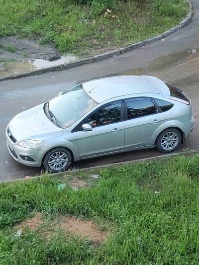 A silver Ford Focus hatchback is parked on a paved surface, likely a street or parking area, with a curb separating it from a patch of overgrown grass and dirt. The car is positioned at an angle, with its front facing towards the lower left of the frame and its rear towards the upper right. The car's windows are tinted, and a portion of the interior is visible, showing at least one person. Puddles of water are present on the paved surface near the curb, suggesting recent rain. The surrounding environment appears to be outdoors, with abundant green vegetation. The scene is consistent with an urban or suburban setting in Belousovo, Russia.
